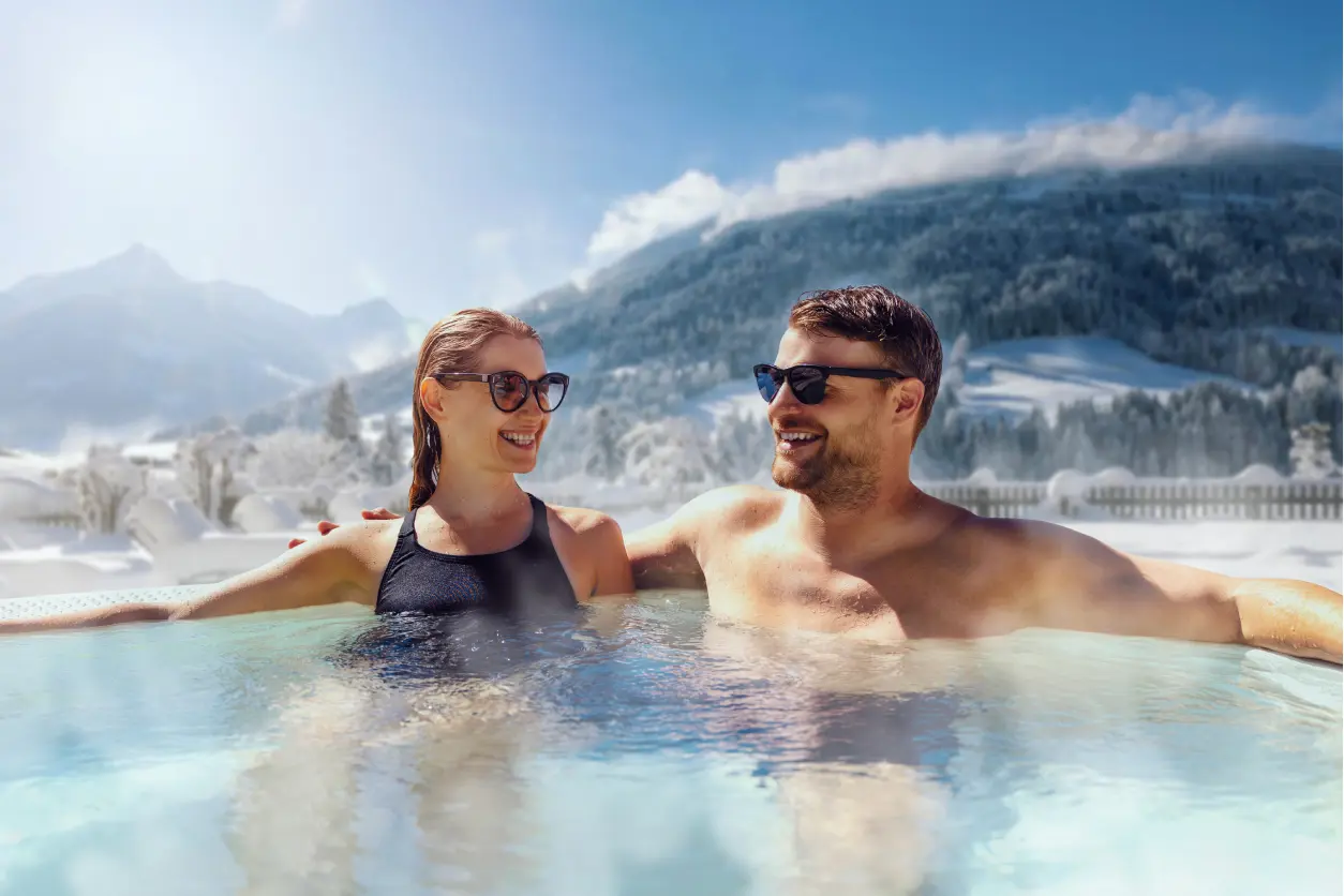 New Project (20) A man and a woman, both wearing sunglasses, relax in an outdoor hot tub with snow-covered mountains in the background.